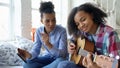 Mixed race young woman with tablet computer sitting on bed teaching her teenage sister to play acoustic guitar at home Royalty Free Stock Photo