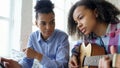 Mixed race young woman with tablet computer sitting on bed teaching her teenage sister to play acoustic guitar at home Royalty Free Stock Photo