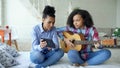 Mixed race young woman with tablet computer sitting on bed teaching her teenage sister to play acoustic guitar at home Royalty Free Stock Photo