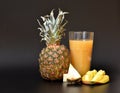 A mixture of fruit juices in a tall faceted glass on a black background, next to pieces of ripe pineapple and mango Royalty Free Stock Photo