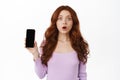 Mobile advertisement. Excited redhead woman gasp wow, shows smartphone empty screen, stare impressed at camera, special