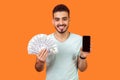 Mobile banking. Portrait of cheerful brunette man holding money and cellphone. indoor studio shot isolated on orange background