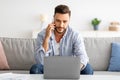 Mobile communication. Young freelancer man talking on cellphone and working on laptop at home on sofa Royalty Free Stock Photo