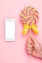 Mobile phone and colorful lolipop with pink, yellow and white spiral in hand on pink background