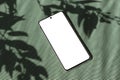 Mobile phone with empty white screen mockup on green textured background with natural floral sunlight shadows, spring