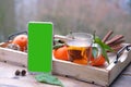 Mobile phone with green screen, glass of hot tea on tray, delicious vitamin tangerines with leaves, cinnamon sticks, pine cones,