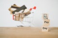 Mobile phone with magnifying glass and shopping cart icon on wooden cube block and pay day word, blurred credit cards in shopping