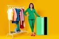 Mobile shopping. Happy lady customer standing near huge credit card and clothing rail on yellow background, full length