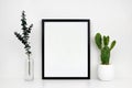 Mock up black frame with cactus and branches on a white shelf or desk against a white wall Royalty Free Stock Photo