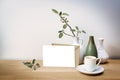 Mock-up of a blank white greeting card with a natural brown envelope, three vases with a sage leaf branch and a coffee cup on a Royalty Free Stock Photo