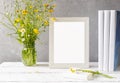 Mock up creation. White frame on white wooden table with grey concrete background, books, notebook and wild flowers Royalty Free Stock Photo