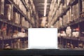 Mock up of modern computer or laptop with blank screen on table with Blurred boxes on rows of shelves in warehouse