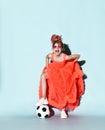 Mocking girl with colorful dreadlocks in bright coral dress, sneakers and sunglasses stands with soccer ball under foot