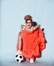 Mocking girl with dreadlocks in sneakers stands with soccer ball under foot holding lifting fur hem of her bright coral dress