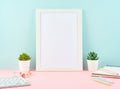 Mockup with blank white frame, alarm, notepad, keyboard on pink table against blue wall with copy space. Modern bright office