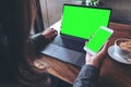 Mockup image of business woman holding mobile phone with blank green screen while using laptop on wooden table Royalty Free Stock Photo