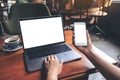 Business woman holding mobile phone with blank white screen while using laptop on wooden table in cafe Royalty Free Stock Photo