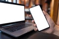 Mockup image of hand holding black mobile phone with blank white screen and black laptop at coffee shop,white blank screen for Royalty Free Stock Photo