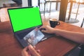 Mockup image of a hand holding credit cards while using and typing on laptop with blank green screen and coffee cup on wooden tabl Royalty Free Stock Photo