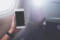 A hand holding and looking at white smart phone with black screen next to an airplane window with clouds and sky background Royalty Free Stock Photo