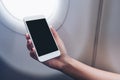 Mockup image of a hand holding and looking at white smart phone with blank desktop screen next to an airplane window Royalty Free Stock Photo