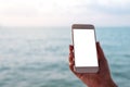 Mockup image of a hand holding and showing white mobile phone with blank black desktop screen in front of the sea and blue sky Royalty Free Stock Photo