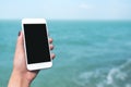 A hand holding and showing white mobile phone with blank desktop screen in front of the sea and blue sky background Royalty Free Stock Photo