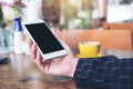 Mockup image of a hand holding white mobile phone with blank black desktop screen and yellow coffee cup on wooden table Royalty Free Stock Photo