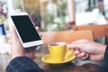 Mockup image of a hand holding white mobile phone with blank black desktop screen and yellow coffee cup on wooden table