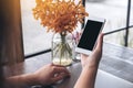 Mockup image of a hand holding white mobile phone with blank black screen with laptop and flower vase on vintage wood table Royalty Free Stock Photo