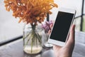 Mockup image of a hand holding white mobile phone with blank black screen with laptop and flower vase on vintage wood table Royalty Free Stock Photo