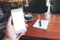 Hand holding white mobile phone with blank desktop screen with coffee cup and notebook on wooden table in cafe Royalty Free Stock Photo