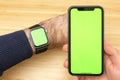 Mockup image of hands holding black mobile phone with green screen and electronic smart watch on wrist . Office table on the. Royalty Free Stock Photo