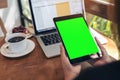 Mockup image of hands holding black tablet pc with blank green screen , laptop , coffee cup and cake on wooden table Royalty Free Stock Photo