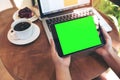 Mockup image of hands holding black tablet pc with blank green screen , laptop , coffee cup and cake on wooden table Royalty Free Stock Photo