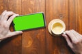 Mockup image of hands holding mobile phone with blank green screen with coffee cup on wooden table. Top view Royalty Free Stock Photo