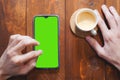 Mockup image of hands holding mobile phone with blank green screen with coffee cup on wooden table. Top view Royalty Free Stock Photo