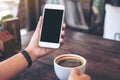 Mockup image of hands holding white mobile phone with blank black desktop screen and a coffee cup on wooden table Royalty Free Stock Photo