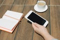 Mockup image of hands holding white mobile phone with blank black desktop screen with notebook and coffee cup on wooden table in Royalty Free Stock Photo