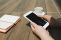 Mockup image of hands holding white mobile phone with blank black desktop screen with notebook and coffee cup on wooden table in Royalty Free Stock Photo