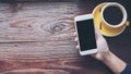 Mockup image of hands holding white mobile phone with blank black screen with yellow hot coffee cups on vintage wooden table Royalty Free Stock Photo