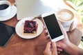 Mockup image of hands holding white smartphone with blank black screen , tablet , laptop , coffee cup and cake on wooden table Royalty Free Stock Photo