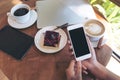 Mockup image of hands holding white smartphone with blank black screen , tablet , laptop , coffee cup and cake on wooden table Royalty Free Stock Photo