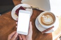 Mockup image of hands holding white smartphone with blank screen , tablet , laptop , coffee cup and cake on wooden table Royalty Free Stock Photo