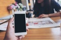 Mockup image of a man`s hand holding white mobile phone with blank black screen in modern cafe and blur woman reading newspaper