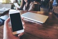 Mockup image of a man`s hand holding white mobile phone with blank black screen in vintage cafe with laptop on wooden table Royalty Free Stock Photo