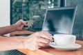 Mockup image of man`s hands holding mobile phone with blank black screen and laptop and a coffee cup on wooden table Royalty Free Stock Photo