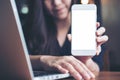 Mockup image of a smiley Asian beautiful woman holding and showing white mobile phone with black screen while using laptop on wood