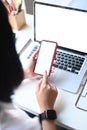 Woman holding smart phone with blank desktop screen while sitting in front of her laptop. Royalty Free Stock Photo