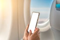 Mockup image of a woman holding at smart phone with blank white screen next to an airplane window with clouds and sky background Royalty Free Stock Photo